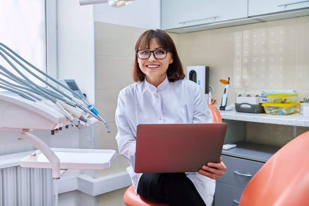 A smiling orthodontist in a clinic holding a laptop, sitting beside dental equipment.