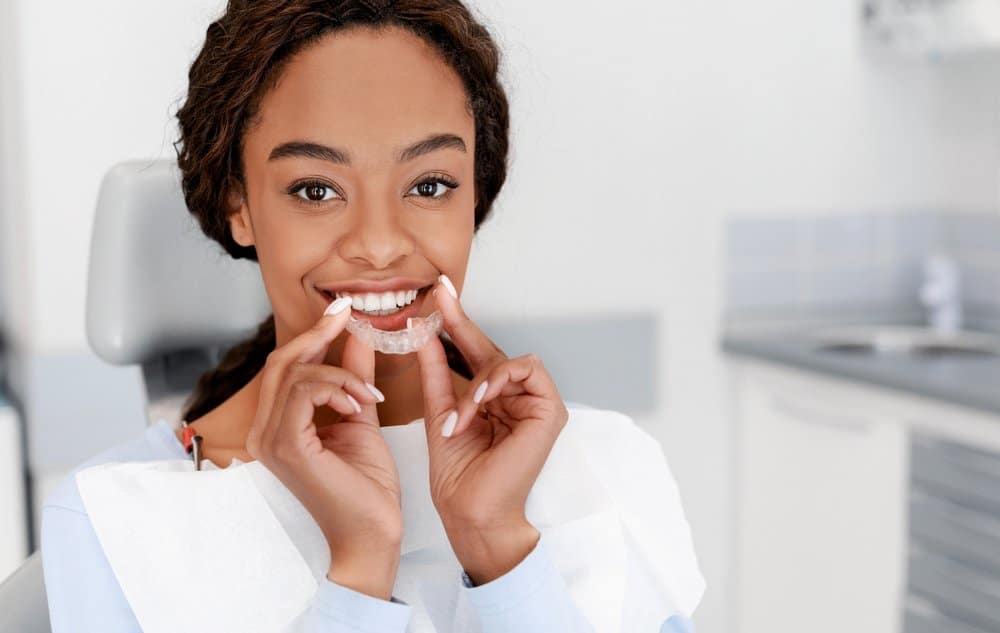 A smiling woman at the orthodontist holding a clear aligner during teeth straightening treatment.